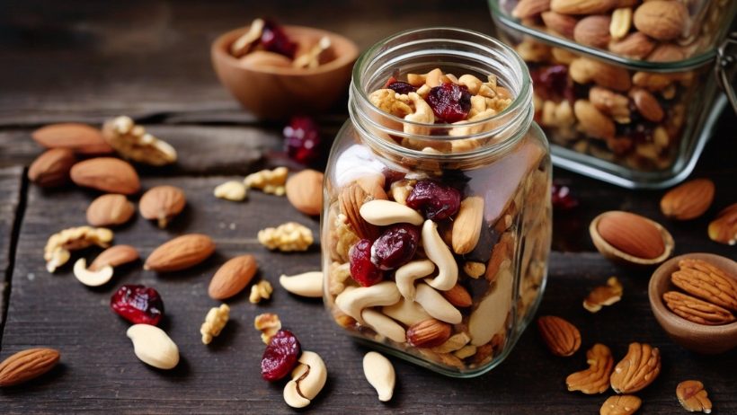 Ein Glas gefüllt mit gemischten Nüssen und getrockneten Cranberries auf einem Holztisch, mit weiteren gemischten Nüssen verstreut herum und einem weiteren Glas und kleinen Holzschüsseln im Hintergrund – perfekt für alle, die vegane Rezepte ausprobieren möchten.