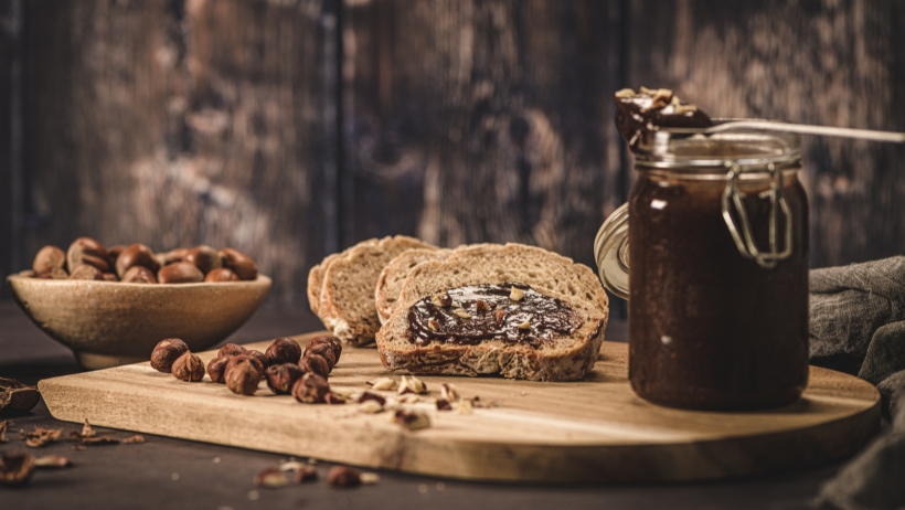 Brotscheiben mit veganem Schokoaufstrich auf einem Holzbrett, ein Glas veganen Schokoaufstrich mit Löffel und eine Schale mit Haselnüssen. Perfekt für Anfänger auf der Suche nach einfachen veganen Rezepten.