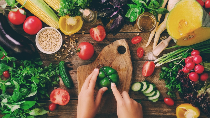 Hände schneiden eine Paprika auf einem Holzschneidebrett, umgeben von verschiedenen frischen Gemüsesorten, darunter Tomaten, Basilikum, Kürbis, Gurken und Radieschen – perfekt für Anfängerrezepte in der veganen Küche.